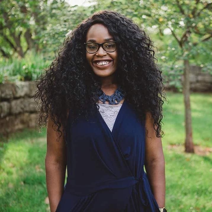 Andrea Ambam wears blue top and glasses and smiles before a tree and a stone wall